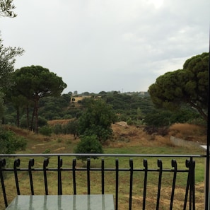 Un balcón con una mesa de vidrio y barandilla metálica, con vistas a un paisaje con árboles y una vista distante de edificios.