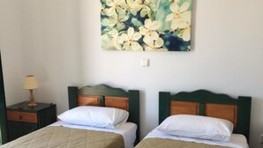 A hotel room with two beds, each with a quilted bedspread and white pillows. There is a painting of flowers on the wall above the beds. - Koukounaries Studios (Kefalonia)