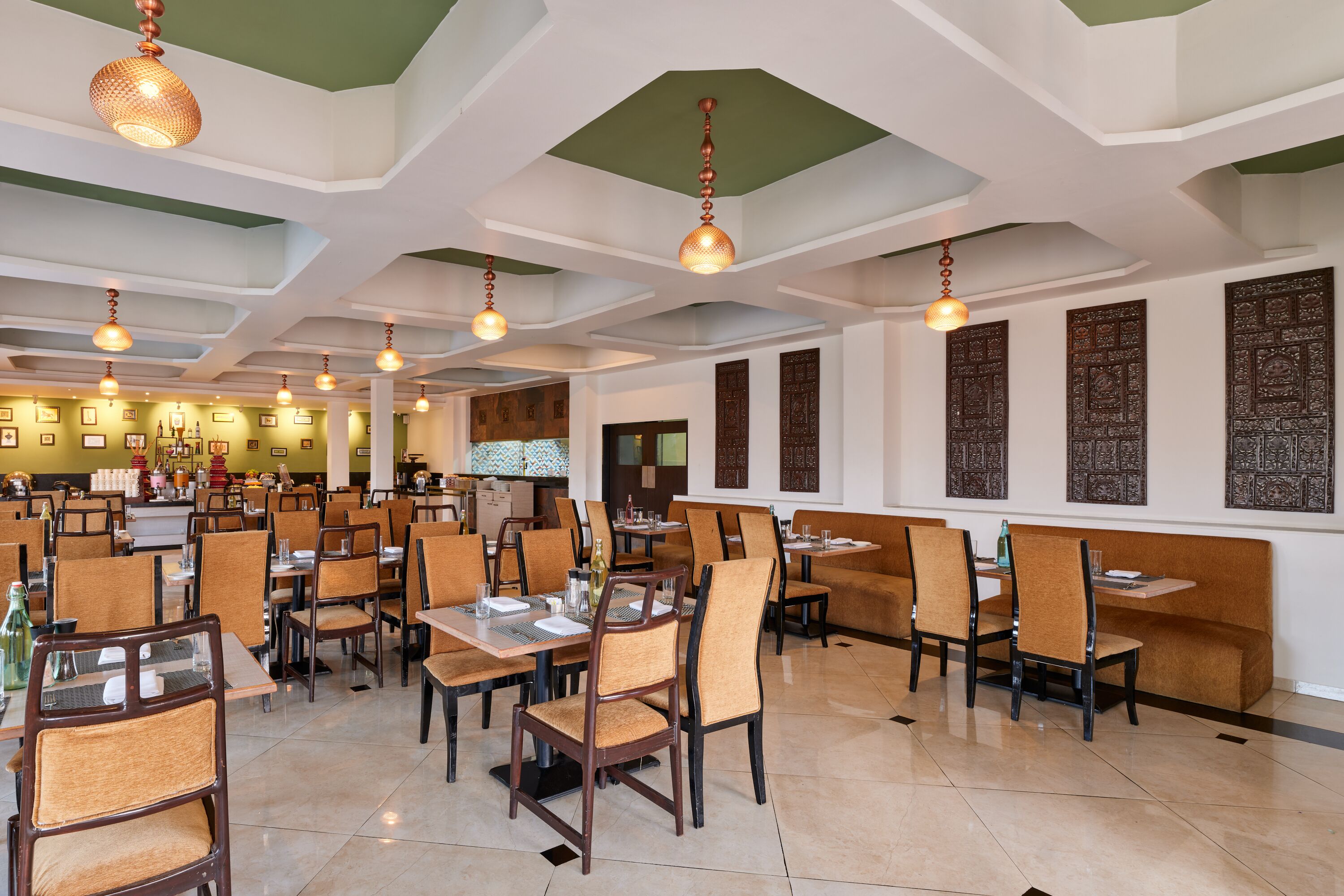 A spacious restaurant with high ceilings, hanging pendant lights, and traditional wall decor. Tables are neatly arranged with chairs, and the floor is tiled.