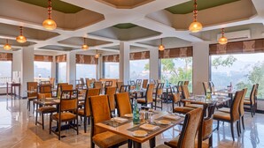 Un restaurante amplio con varias mesas y sillas, ventanas grandes con cortinas y lámparas colgantes.