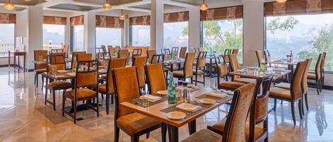 A spacious restaurant with multiple tables and chairs, large windows with curtains, and hanging pendant lights.