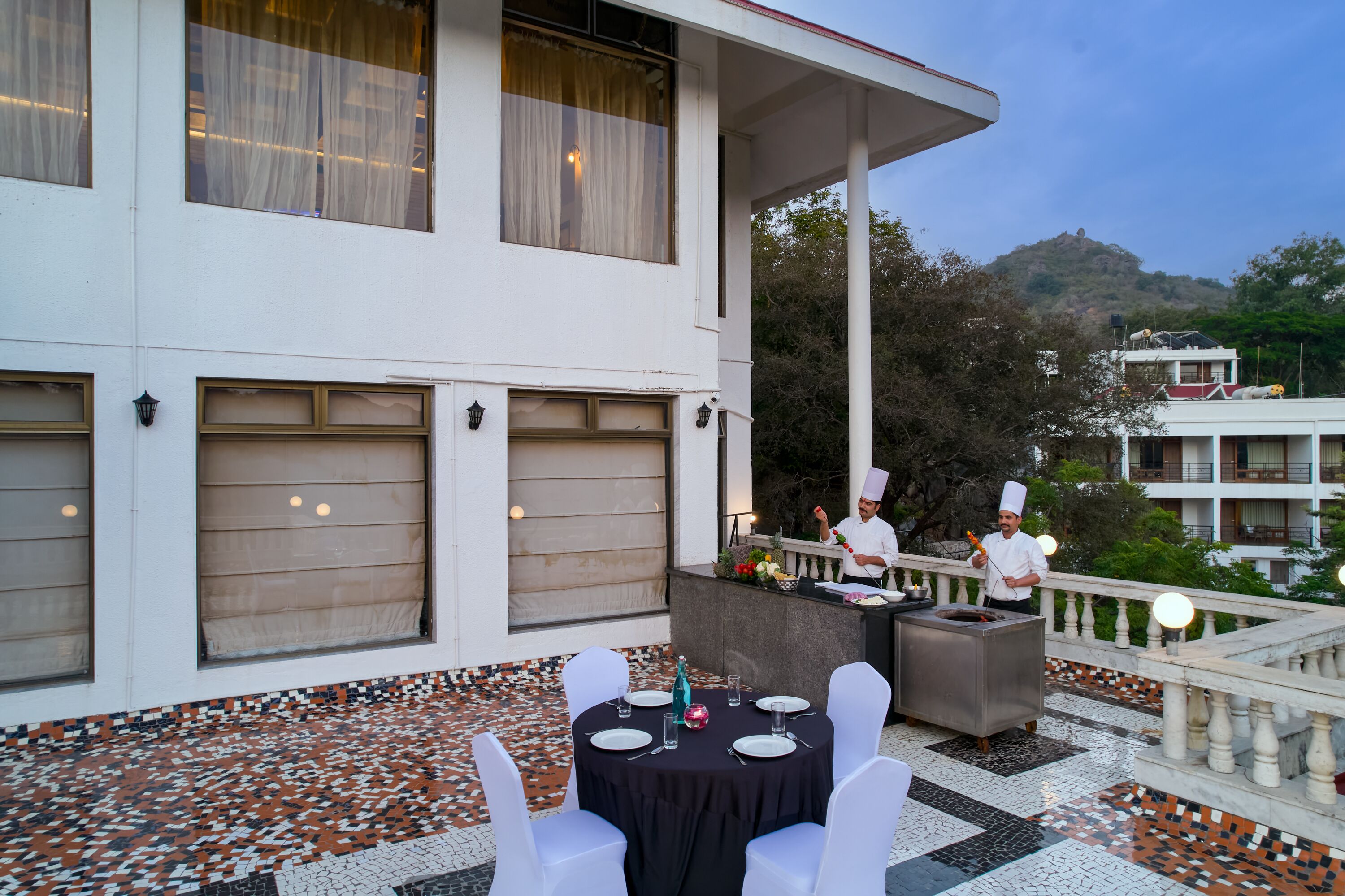An outdoor dining area with a table set for two, a chef preparing food, and a view of a hill in the background.