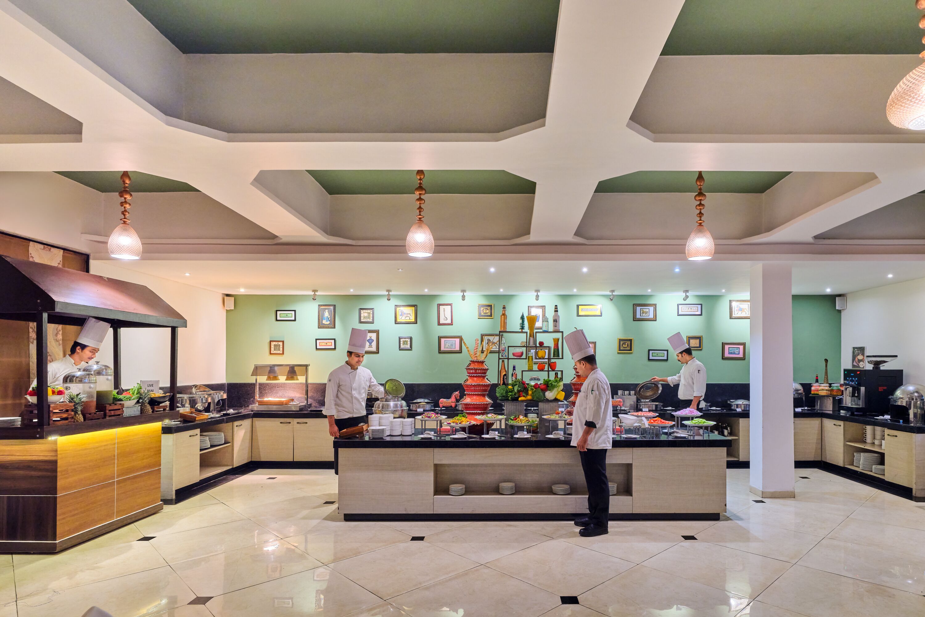 A modern kitchen with chefs preparing food, a buffet setup, and a green wall with framed pictures.
