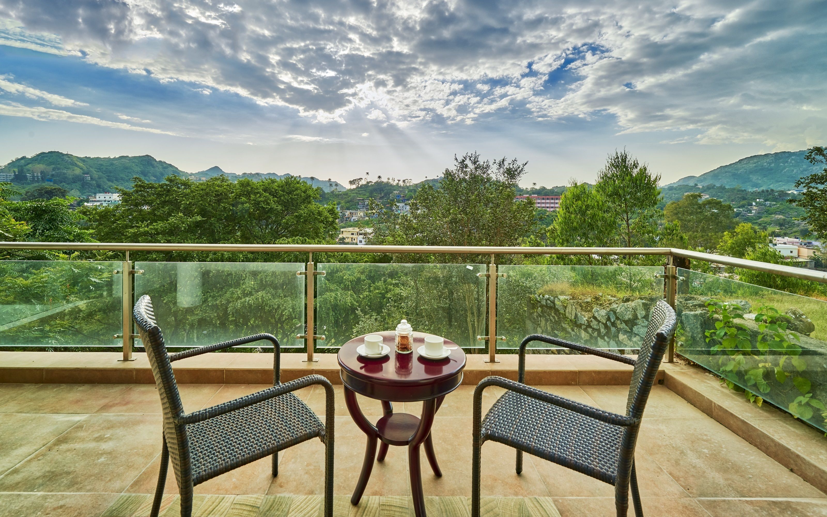 A balcony with a round table and two chairs, overlooking a scenic landscape with mountains and trees.