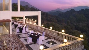 Un área de comedor al aire libre con mesas y sillas, barandilla de balcón y montañas al fondo.