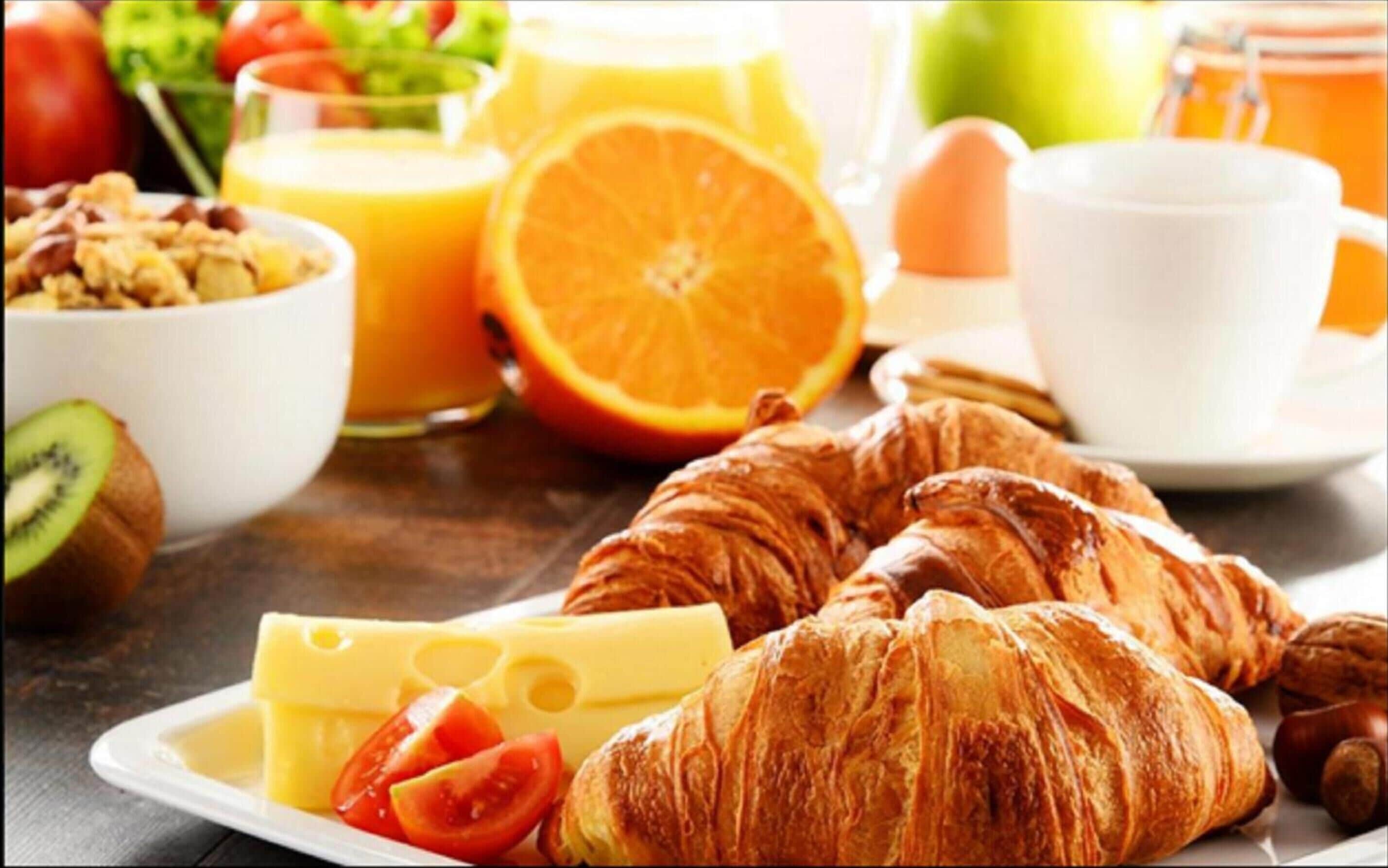 A breakfast setting with croissants, cheese, tomatoes, a glass of orange juice, and a bowl of granola.