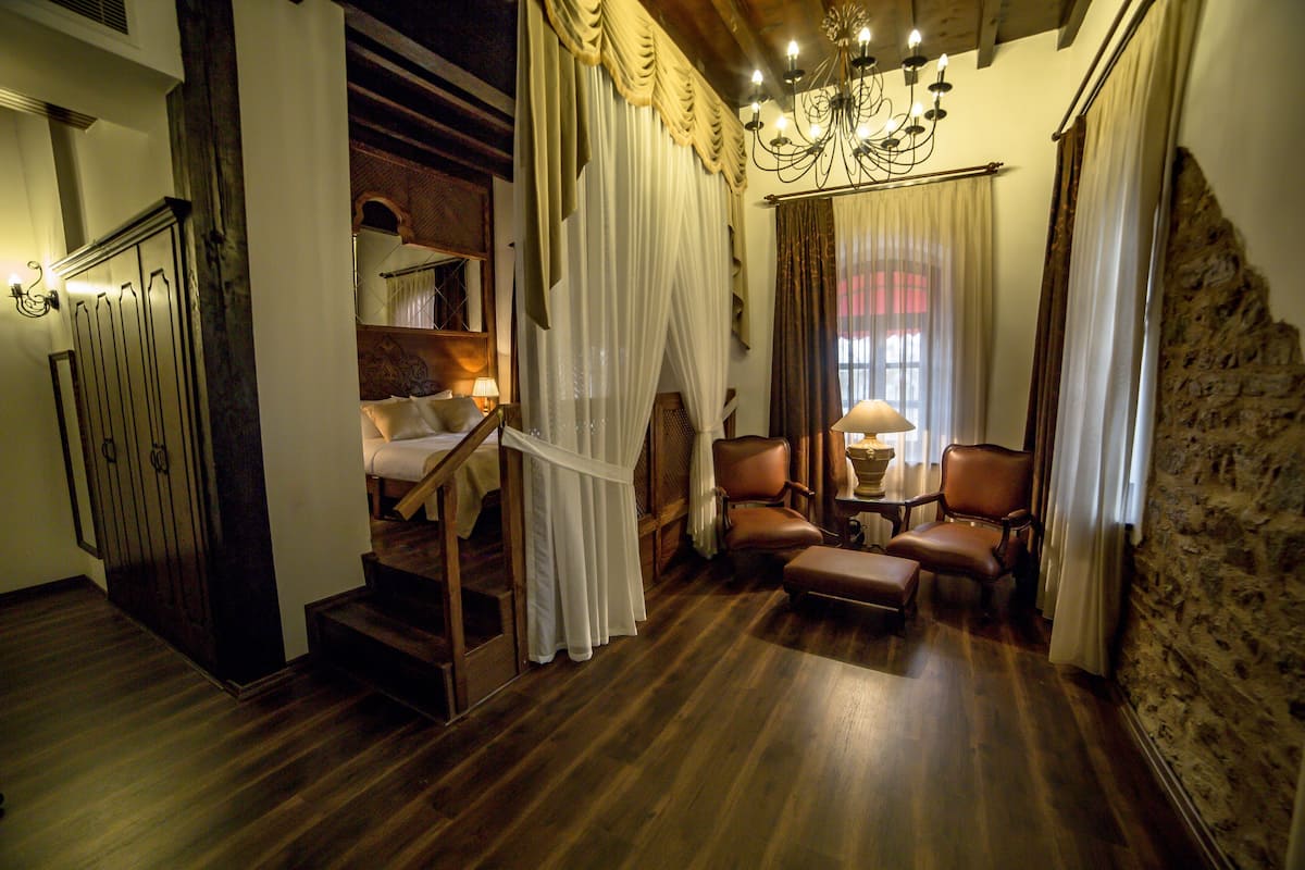 A room with a wooden staircase, a bed, two chairs, a table, and a chandelier.