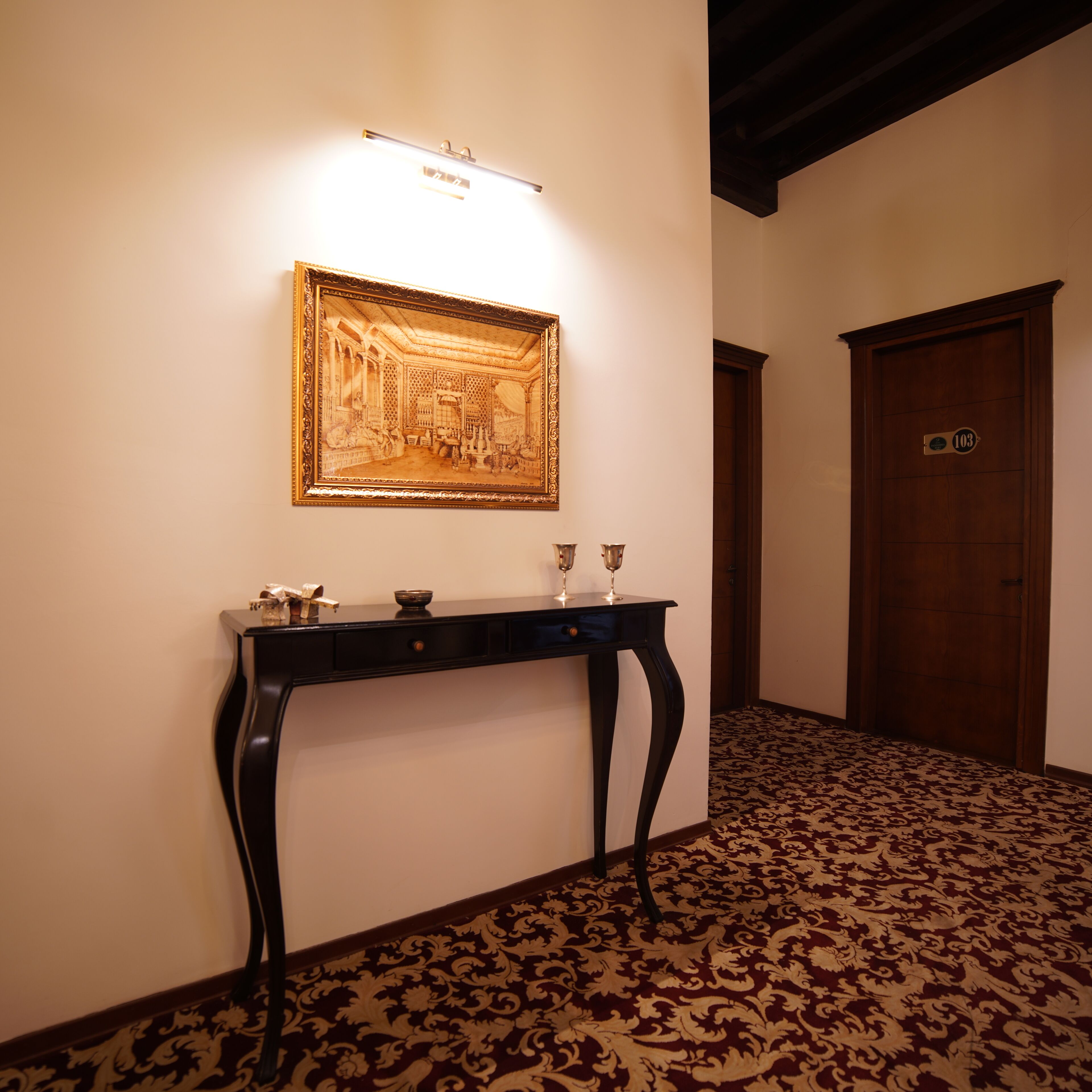 A hallway with a framed artwork on the wall, a dark wooden table with two glasses, and a patterned carpet.