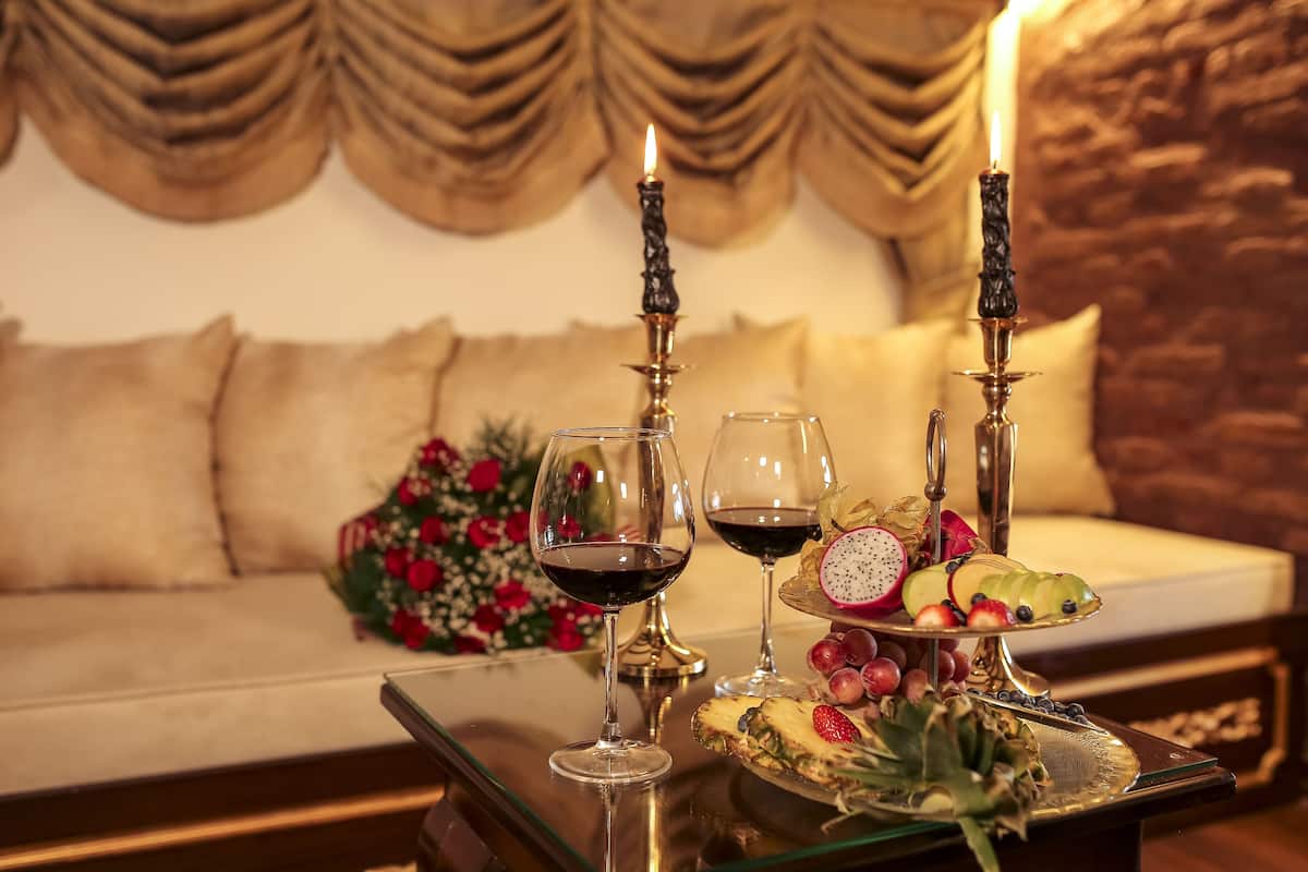 A table with two wine glasses, a fruit platter, and two candles in a cozy room with a sofa and a decorative wall.