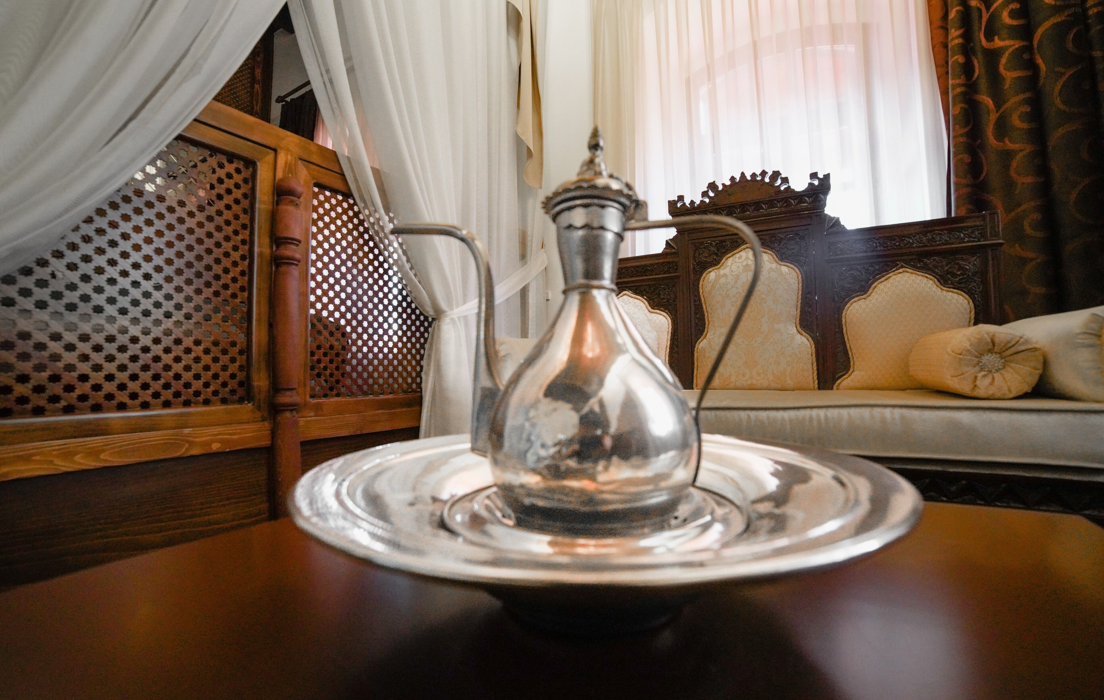 Una tetera metálica tradicional sobre una bandeja, una silla de madera, y un sofá con cojines en una habitación con cortinas estampadas y un cabecero de madera.