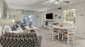 A spacious living room with a white sofa, patterned armchair, and dining table with white chairs. A ceiling fan and TV are mounted on the wall.