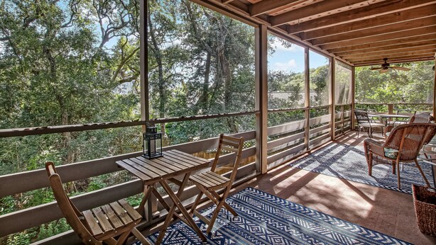 A covered outdoor area with wooden furniture, a patterned rug, and large windows offering a view of trees.