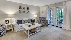 A living room with a white sofa, patterned armchair, and a coffee table with a lamp. There are framed pictures on the wall and a view of the outdoors through a glass door.