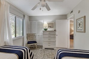 Une chambre avec deux lits, un ventilateur de plafond, une commode, une chaise et une porte donnant sur une autre pièce.