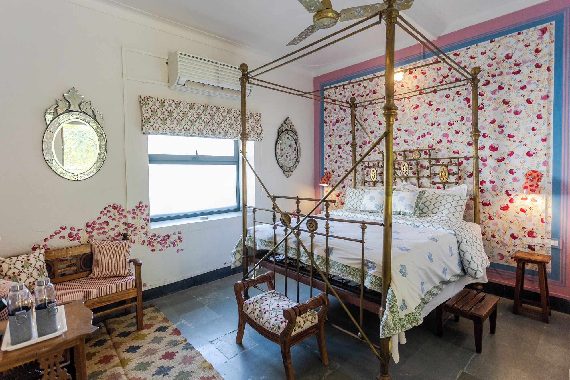 Una cama con dosel, papel tapiz floral, una silla de madera, una mesita con botellas y una ventana con cortina.