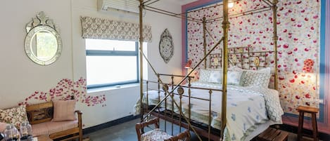 Una cama con dosel, papel tapiz floral, una silla de madera, una mesita con botellas y una ventana con cortina.