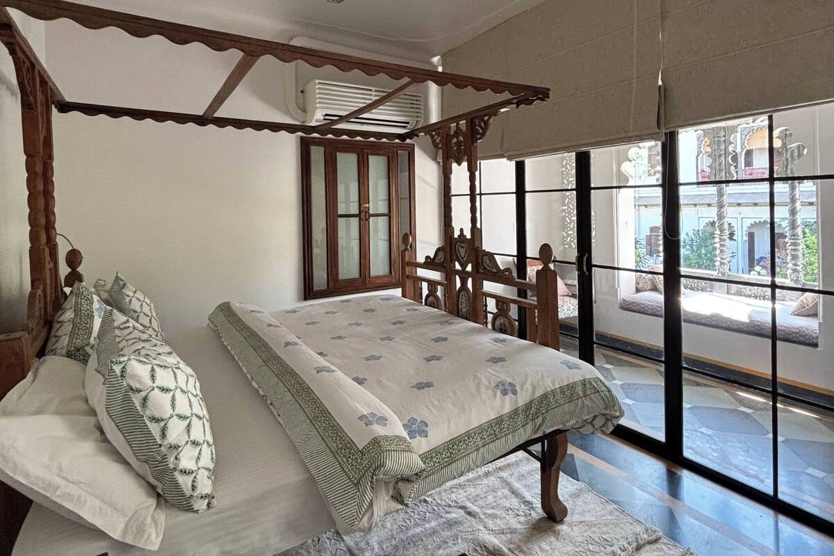 A bedroom with a wooden four-poster bed, a small wooden stool, and a large window providing a view of the outside.