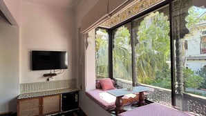 A room with a bed, a TV, a ceiling fan, and a window with a view of greenery. - Kintsugi at Rangniwas (Udaipur)
