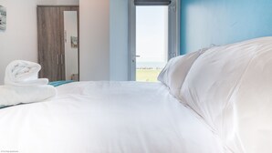 A hotel room with a bed, a window with curtains, a door, and a towel on the bed. - Five Way Apartments (Paignton)