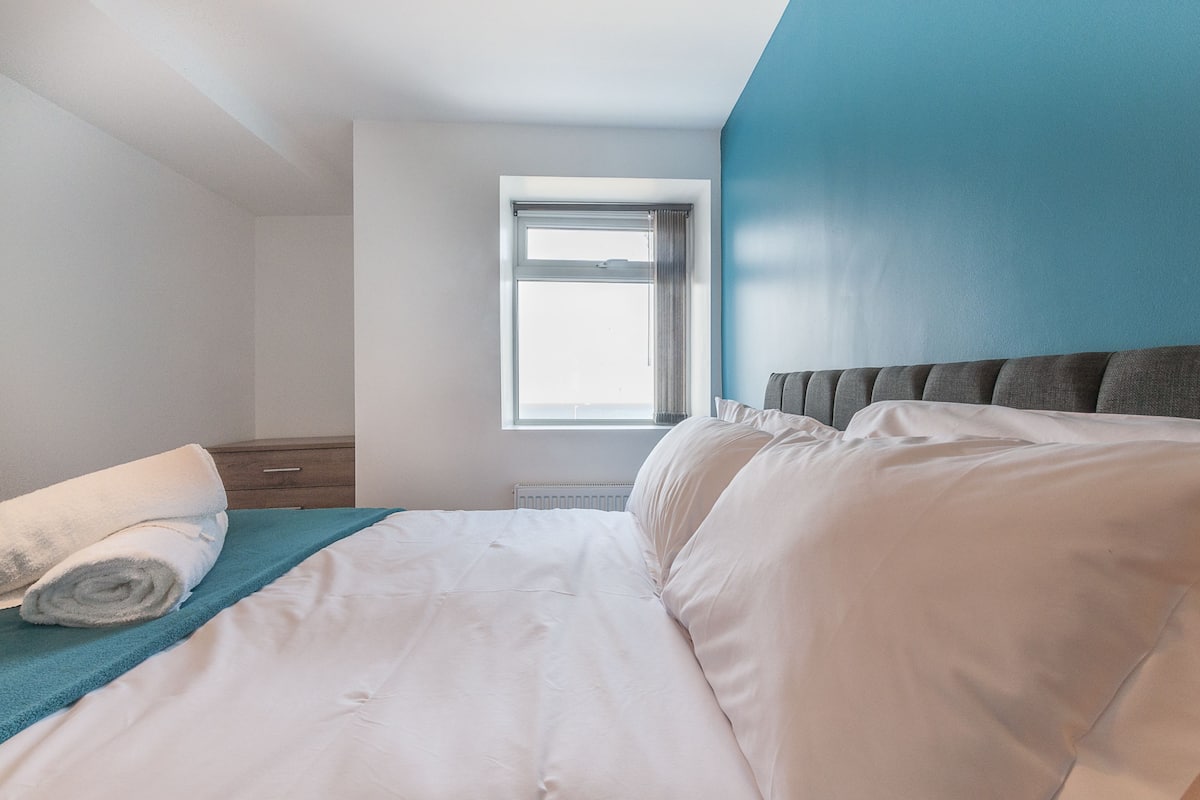 Quarto de hotel com cama, janela, parede azul e mesa de cabeceira de madeira.