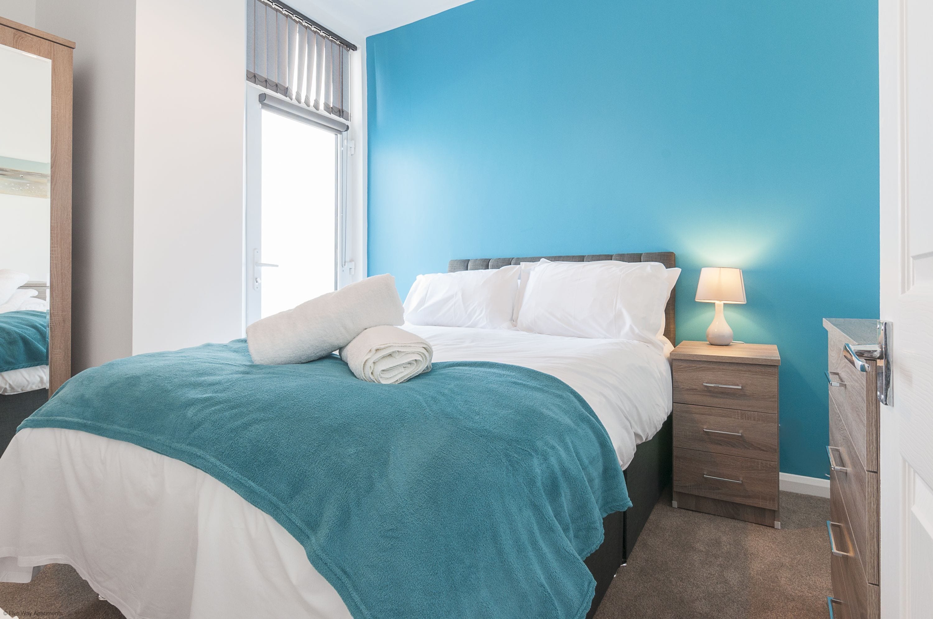 A neatly made bed with a teal blanket, a white pillow, and a rolled towel. There is a bedside table with a lamp and a wooden dresser.