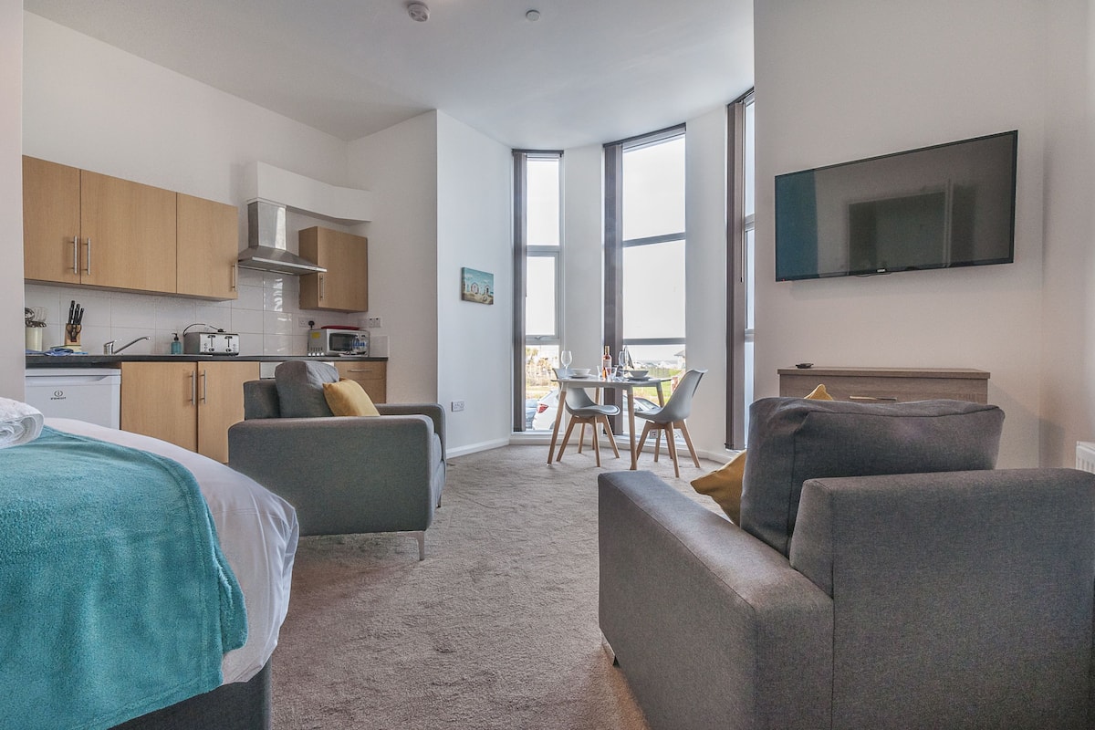 Quarto de hotel moderno com cama, dois sofás, mesa de jantar com cadeiras, cozinha e televisão fixada na parede.