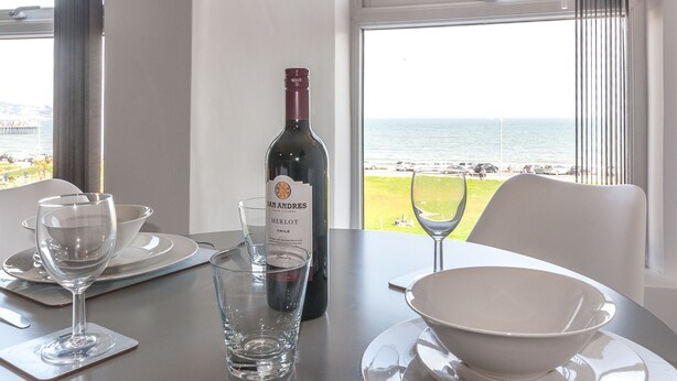 A table with a wine bottle, glasses, and a bowl, overlooking a beach. - Five Way Apartments (Paignton)