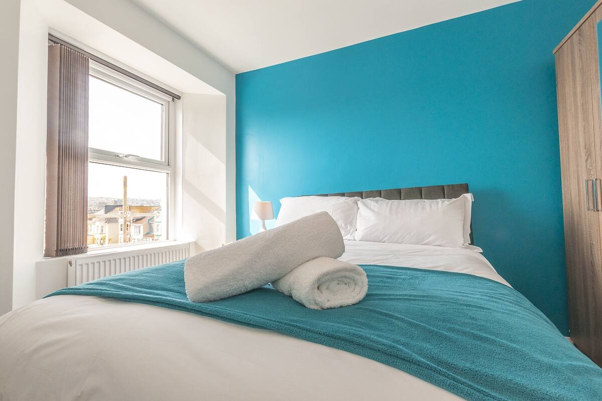 Uma cama bem arrumada com um cobertor azul e uma toalha branca enrolada, uma janela com cortinas e vista para edifícios.