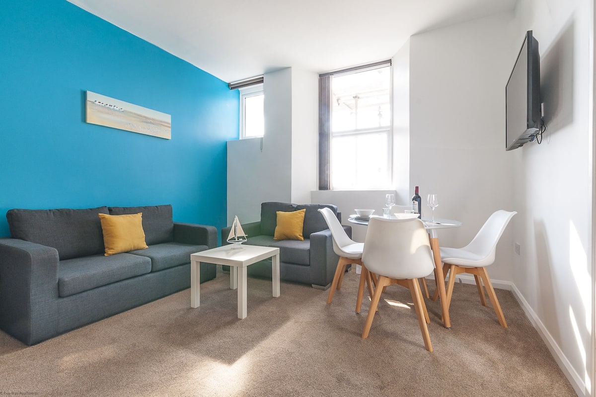 Uma sala de estar moderna com uma parede azul de destaque, um sofá cinza, uma mesinha branca e uma área de jantar com cadeiras brancas.