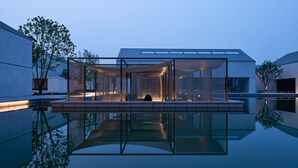 A modern building with a glass facade, a reflective pool in front, and a person sitting on a bench inside. - Alila Wuzhen, Zhejiang (Jiaxing)