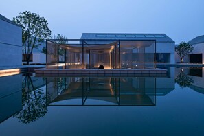 A modern building with a glass facade, a reflective pool in front, and a person sitting on a bench inside. - Alila Wuzhen, Zhejiang (Jiaxing)