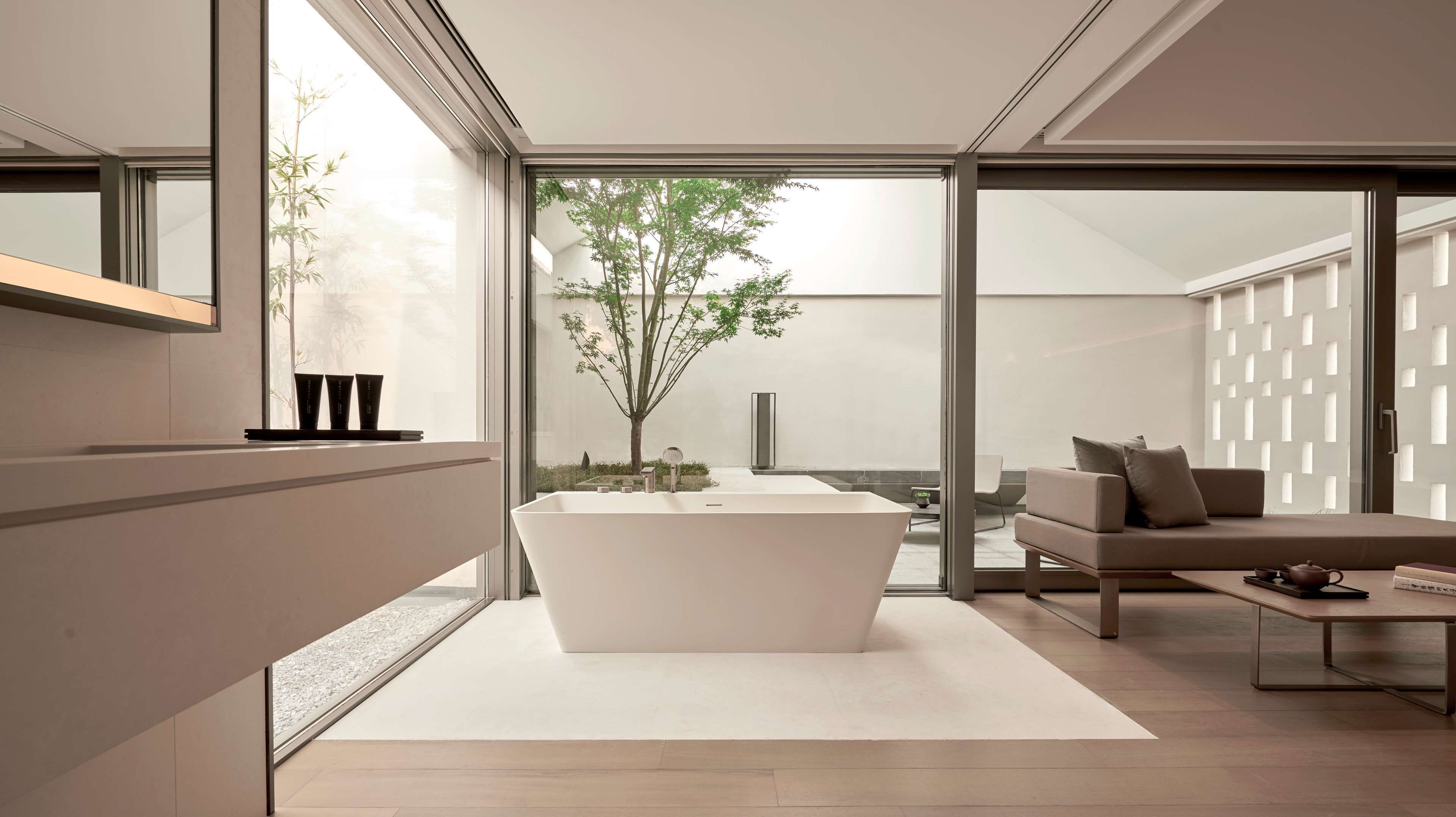 A modern bathroom with a large bathtub, a sink, and a mirror. There is a window with a view of a tree and a balcony.