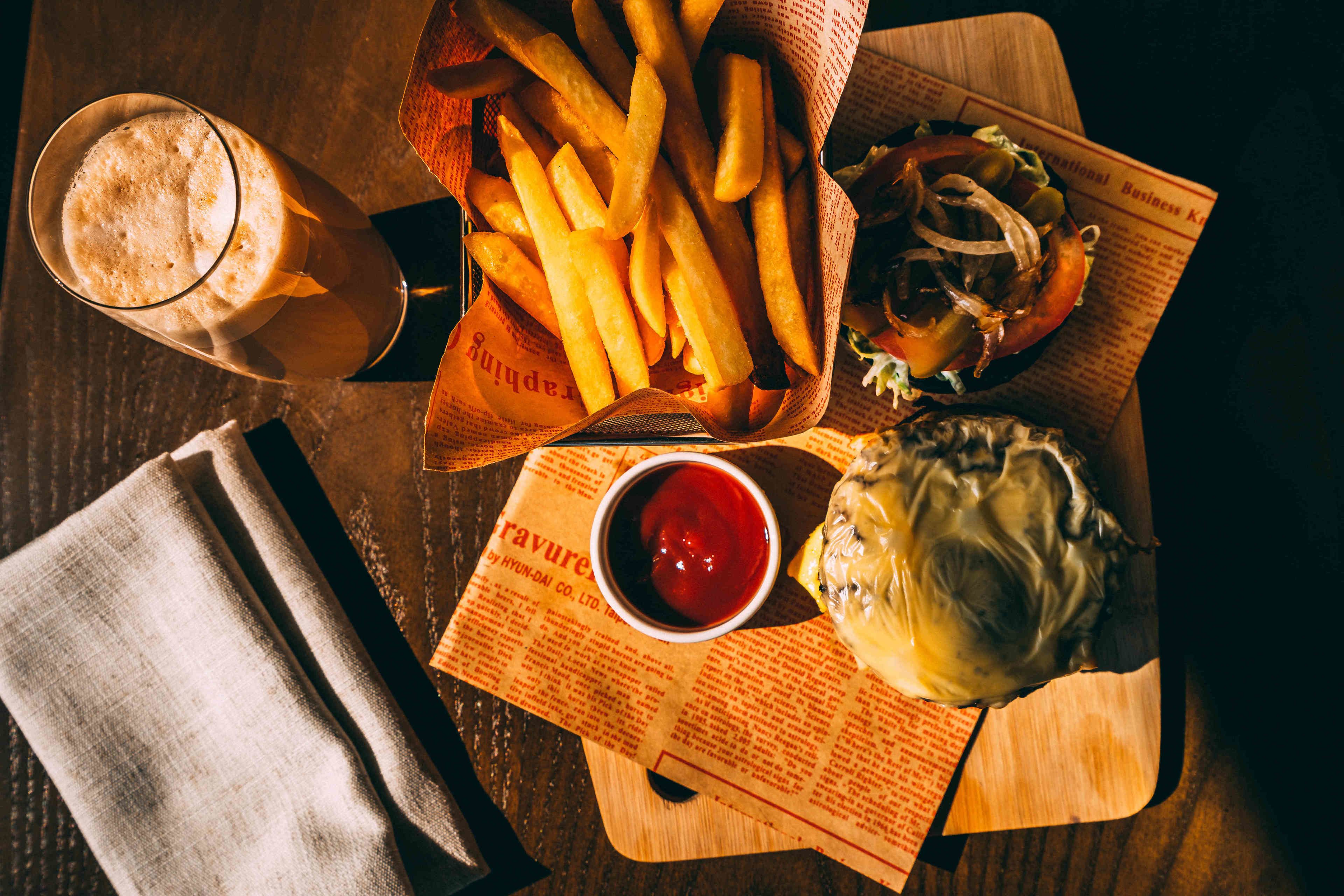 A meal consisting of fries, a burger, and a drink on a wooden table with a newspaper underneath.