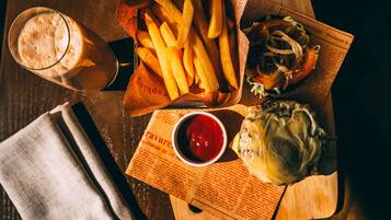A meal consisting of fries, a burger, and a drink on a wooden table with a newspaper underneath.