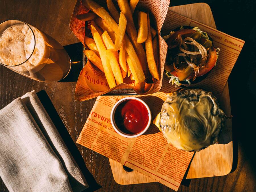 A meal consisting of fries, a burger, and a drink on a wooden table with a newspaper underneath.