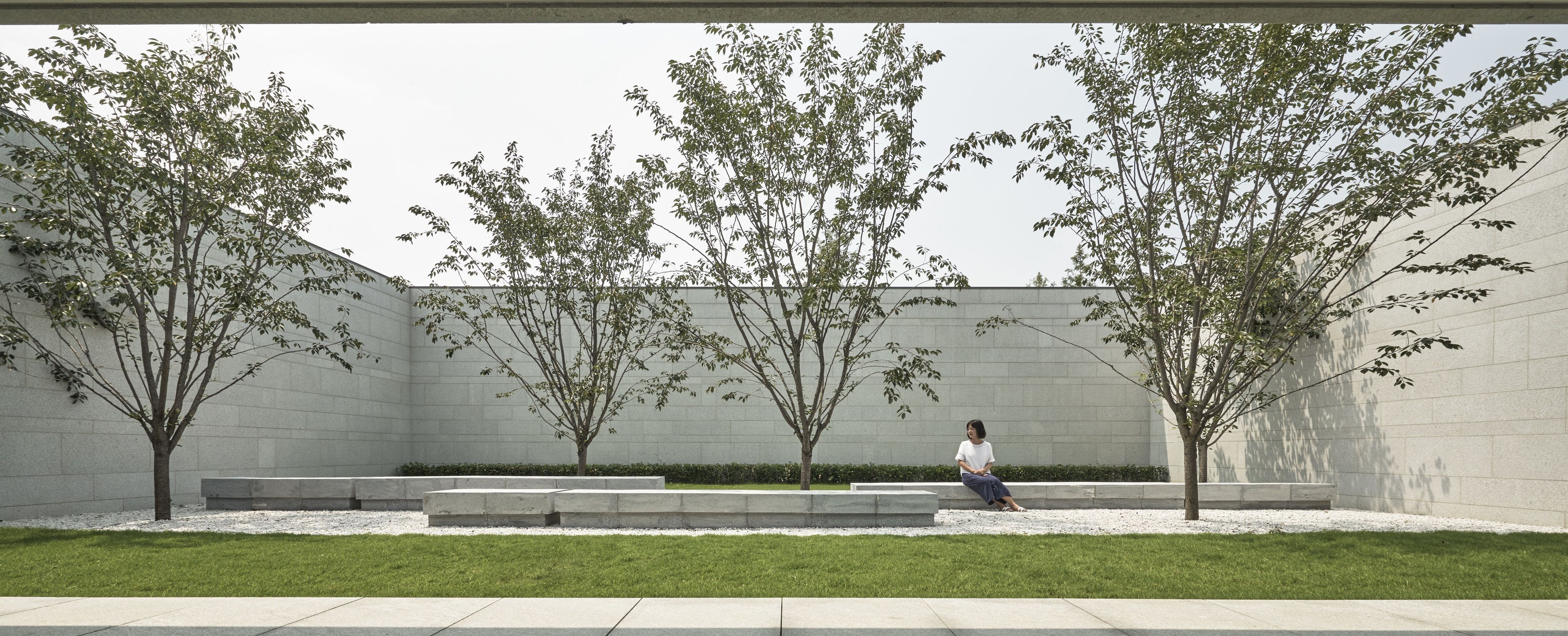 A modern outdoor area with a seating bench, trees, and a building in the background.