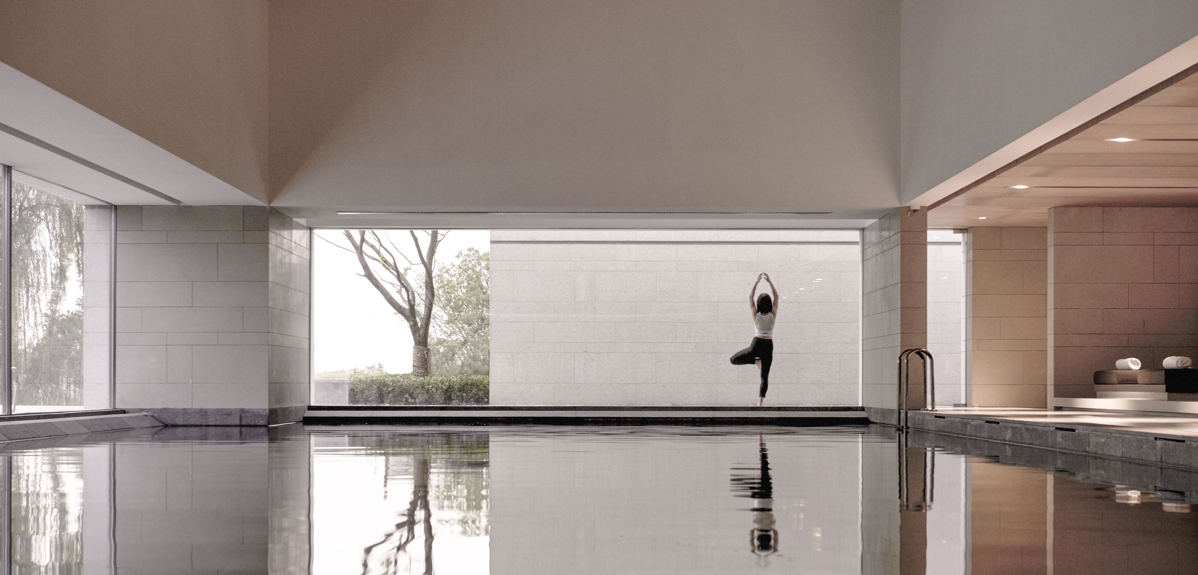 A modern indoor swimming pool with a clear view of trees outside, a tiled wall, and a minimalist design.