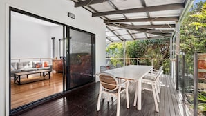 A modern outdoor patio with a wooden deck, a glass door leading to an interior living area, and a covered seating area with a table and chairs.