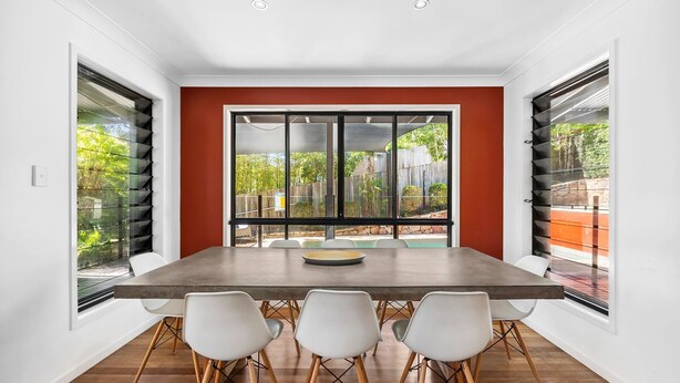 A dining area with a large table, four chairs, and large windows with blinds.