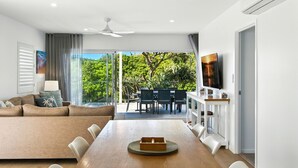 A modern living room with a large sofa, dining table, and chairs. There is a TV mounted on the wall and a ceiling fan. The room has large windows with a view of greenery outside.
