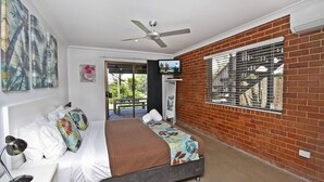 Quarto com cama, ventoinha no teto, televisão e janela com vista para o verde.