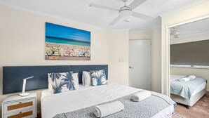 A hotel room with a bed, a nightstand, a ceiling fan, a painting of a beach scene, and a door leading to another room. - Unit 4 'The Rocks', Coolum Beach (Coolum Beach)