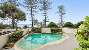 A swimming pool with a pebble edge, surrounded by various trees and a view of the ocean. - Unit 4 'The Rocks', Coolum Beach (Coolum Beach)