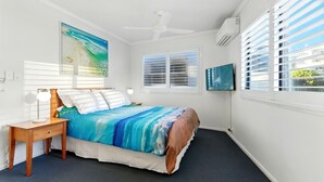 Une chambre à coucher comprenant un lit, une table de chevet, un ventilateur de plafond, un téléviseur et une fenêtre avec des stores.