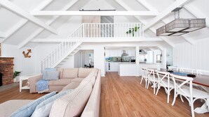 Ein geräumiger Loft mit hoher Decke, einer Holztreppe, einer gemütlichen Wohnzone mit Sofa und Kissen, einem Esstisch mit Stühlen und einer Küchenzeile.