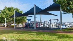 A playground with climbing structures and swings, shaded by large canopies, in a park with a marina in the background.