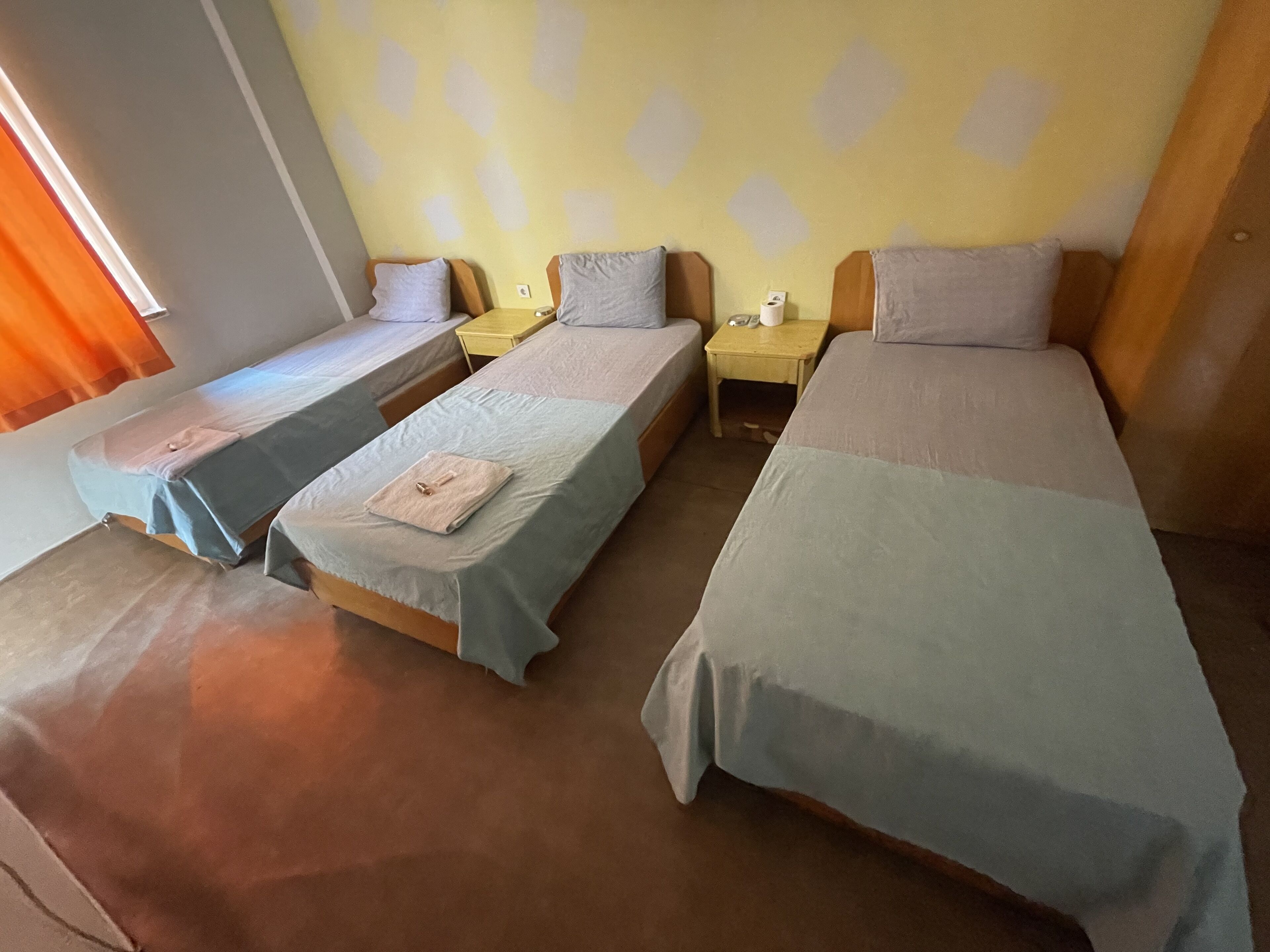 Quarto com três camas individuais, cada uma com cabeceira de madeira e uma pequena mesa de cabeceira. As paredes são pintadas de amarelo com um padrão de losangos, e há uma janela com cortinas laranja no lado esquerdo.
