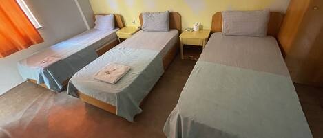 Quarto com três camas individuais, cada uma com cabeceira de madeira e uma pequena mesa de cabeceira. As paredes são pintadas de amarelo com um padrão de losangos, e há uma janela com cortinas laranja no lado esquerdo.