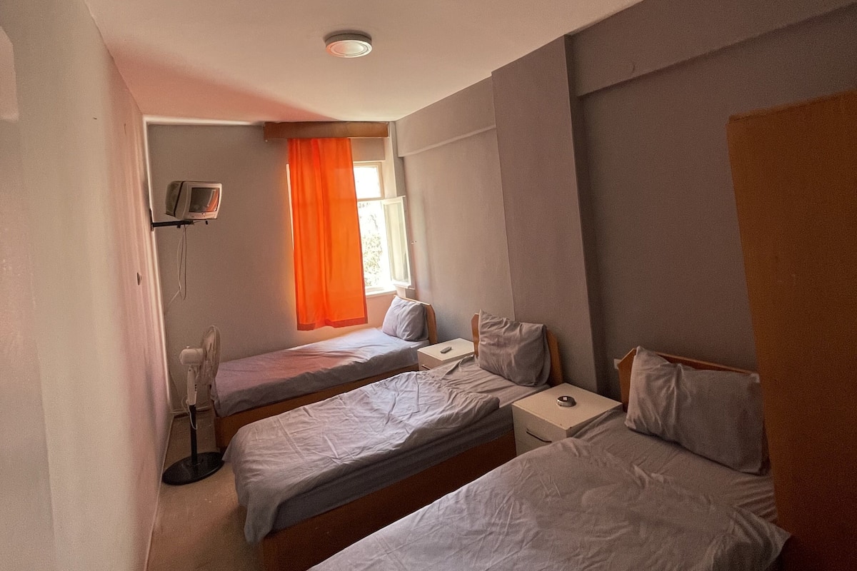 A hotel room with two single beds, a bedside table with a lamp, a window with orange curtains, and a ceiling fan.