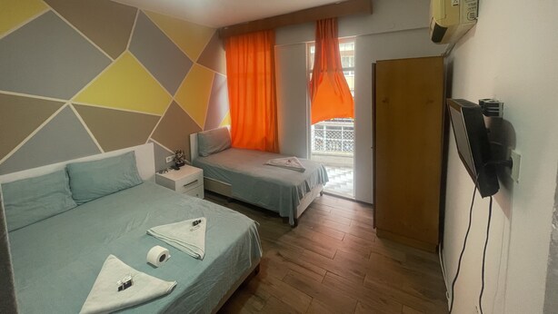 A hotel room with two beds, a small table, a TV, and a window with orange curtains. - Ersoy Ak Pansiyon (Antalya)